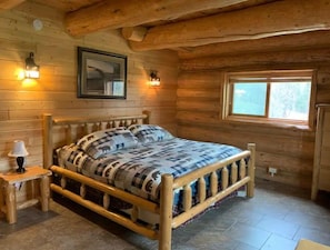 Camera da letto in una baita di legno, con un letto, due lampade a parete, una finestra e un quadro appeso al muro.