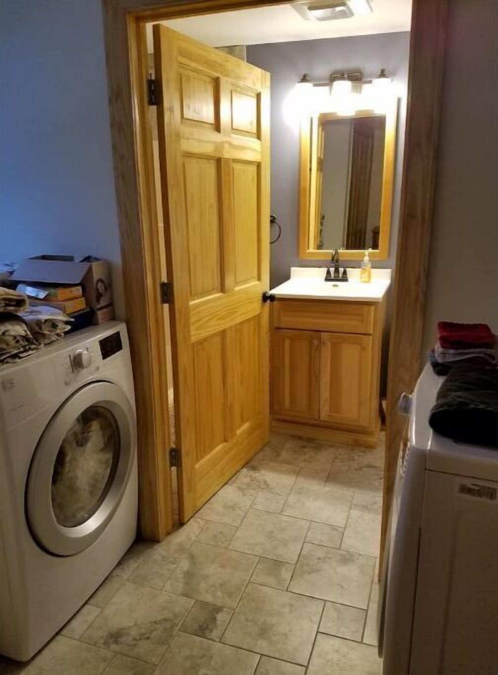 Ein Waschraum mit Waschmaschine, Holztür, Spüle mit Spiegel und einer Arbeitsfläche mit Toilettenartikeln.