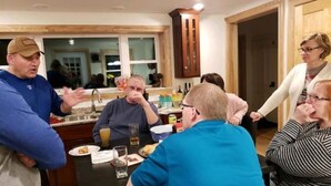 Un groupe de personnes est assis autour d’une table dans une pièce où se trouvent des meubles de rangement en bois et une fenêtre.