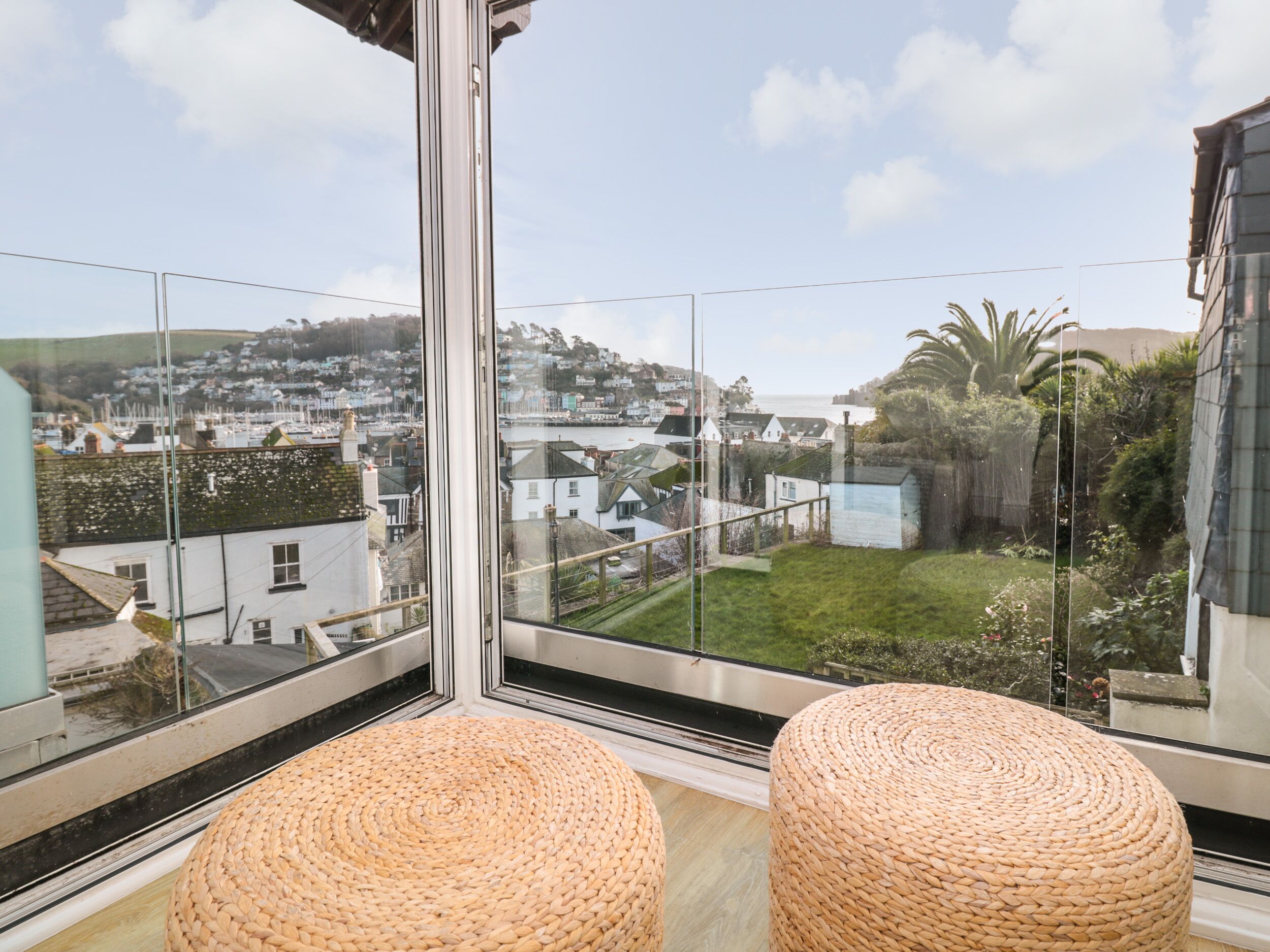 A balcony with wicker ottomans, a glass railing, and a view of a coastal town.