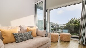 A modern living room with a beige sofa, two ottomans, and a large window offering a view of a coastal town. - Seaview (Dartmouth)