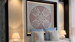 A modern bedroom with a large bed, two bedside tables with lamps, patterned pillows, and a decorative wall piece above the bed. - Aquamarina Pintadera Rossa & SPA (Porto Pino)