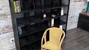Une pièce avec une étagère noire adossée à un mur de briques, contenant des livres, un panneau d’accueil encadré et une petite table avec une lampe.