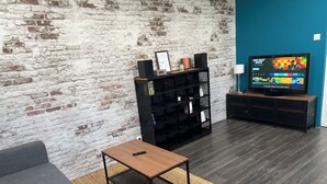 Un salon avec un mur en briques, un canapé gris, une table basse en bois, un meuble de télévision noir et un téléviseur diffusant une chaîne de sport.