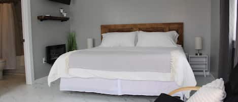 Un dormitorio con cama, televisor montado en la pared, aire acondicionado, mesita de noche con lámpara y una silla con toalla.