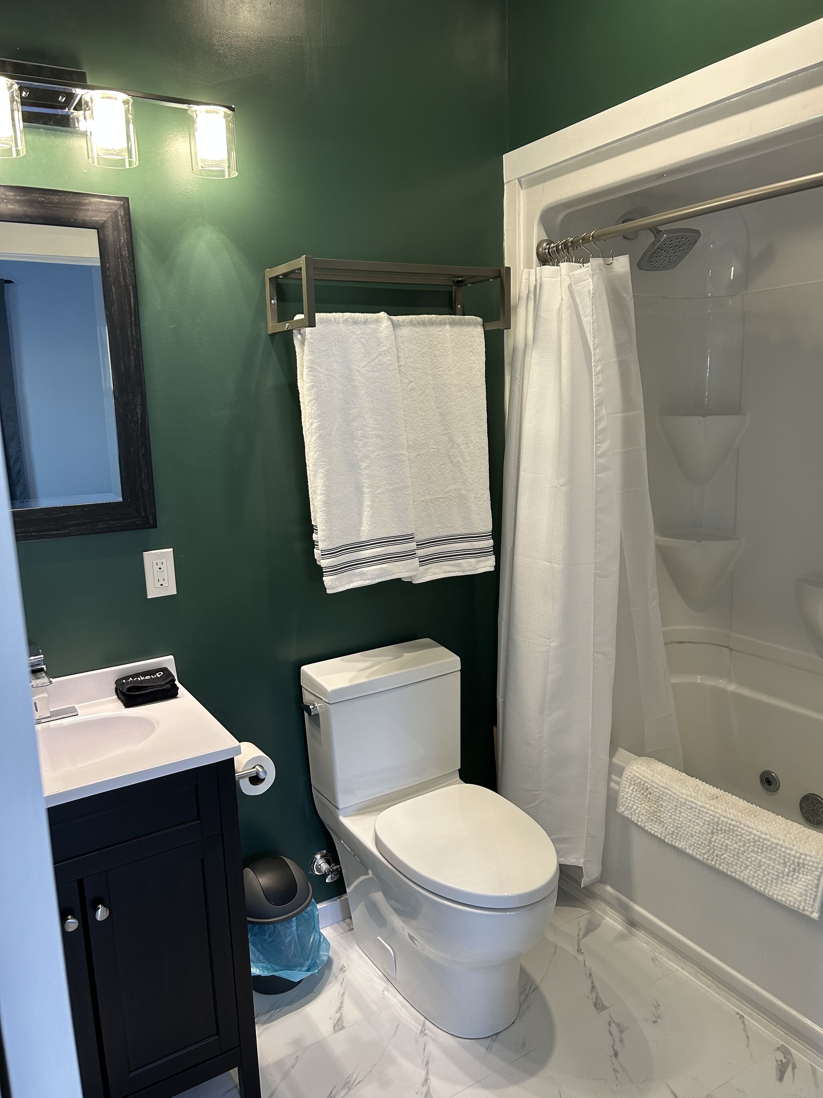 Un bagno con un water bianco, un lavandino con dispenser di sapone nero, una doccia con tenda bianca e una parete verde.