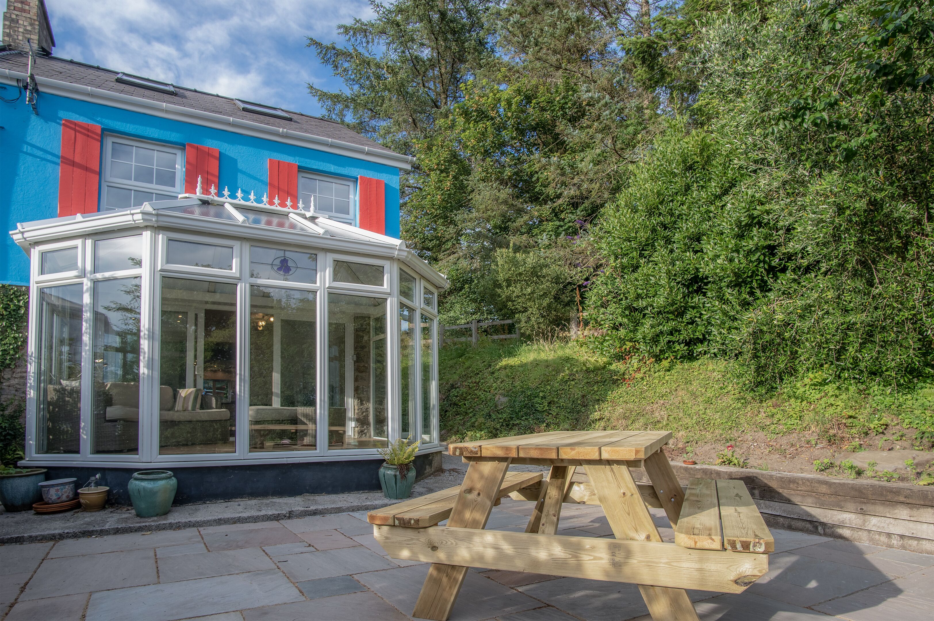 A conservatory with a glass roof, a wooden picnic table, and potted plants.