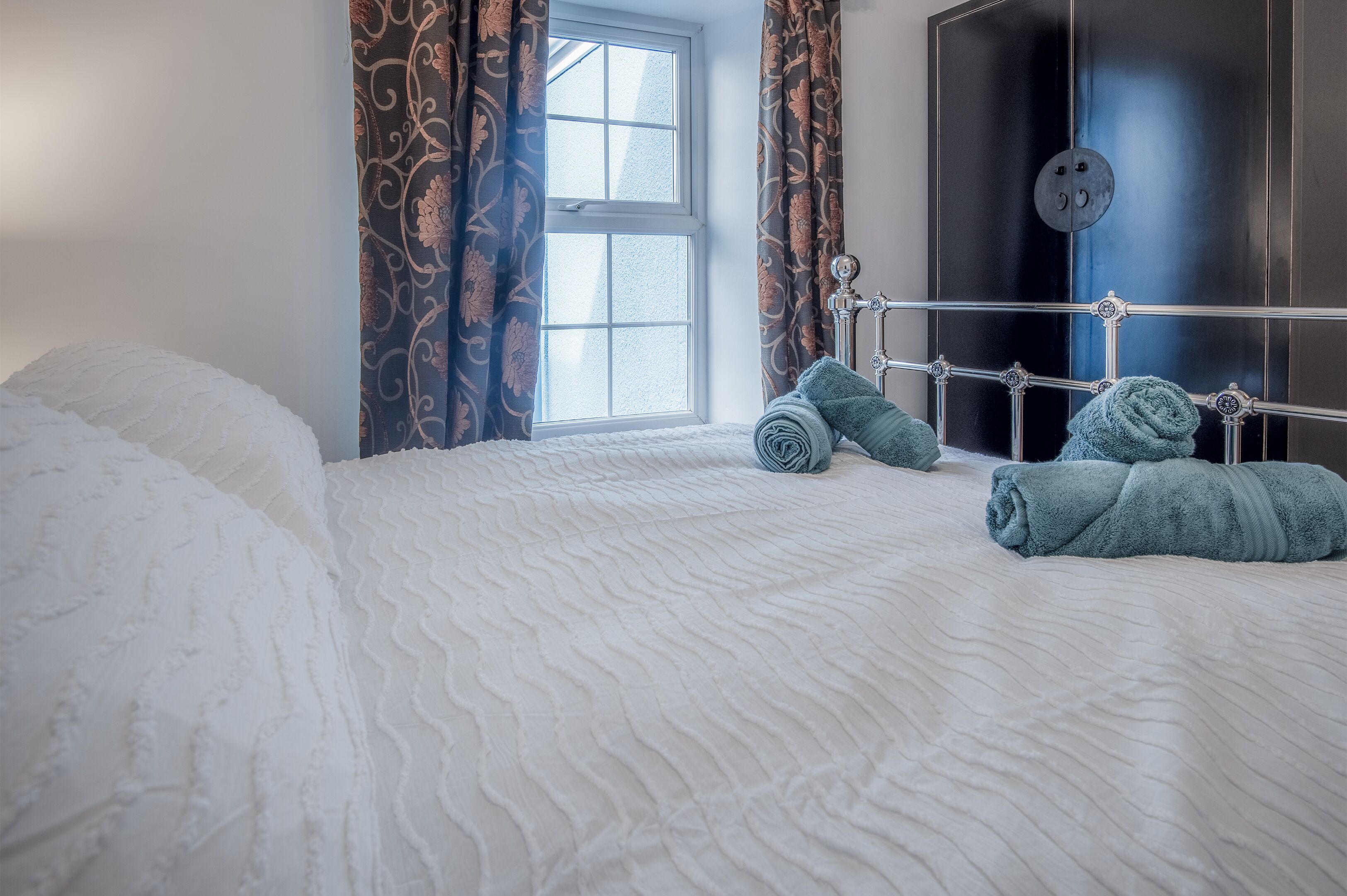 A neatly made bed with white bedding and two rolled towels on a metal headboard.