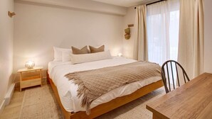 Un dormitorio con una cama de madera, una mesita de noche con lámpara, una mesa de comedor y un ventanal con cortinas.