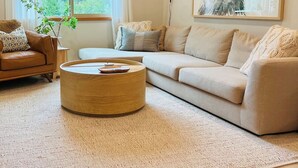 Una sala de estar moderna con un sofá color beige, una mesa de centro redonda de madera y un ventanal con cortinas translúcidas.