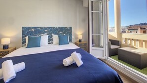A hotel room with a large bed, a patterned headboard, two bedside tables with lamps, and a balcony with a view of the city.
