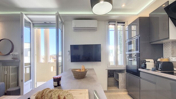 A modern kitchen with a large island, a TV mounted on the wall, and a view of the outside through the open door.