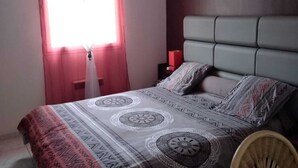 Un dormitorio con una cama, una lámpara colgante roja, un ventanal con cortinas y una silla de madera.