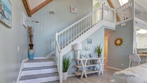 Una escalera con barandillas blancas que conduce a un segundo piso, un comedor con un adorno en la pared con forma de sol y una sala de estar con un sofá de color claro.