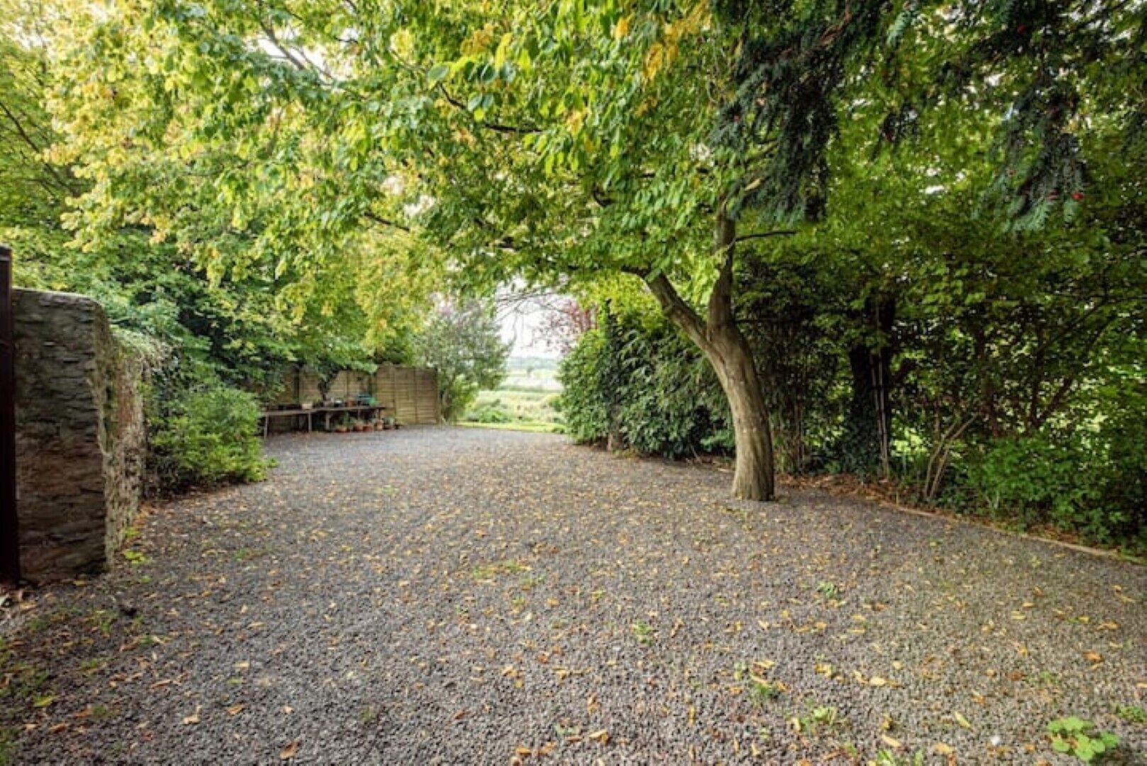 Een grindpad leidt naar een stenen muur, omringd door weelderige begroeiing en bomen.
