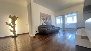 A spacious living room with a wooden floor, a black leather sofa, a white radiator, and a cityscape painting on the wall. - SkyLine (Bari)