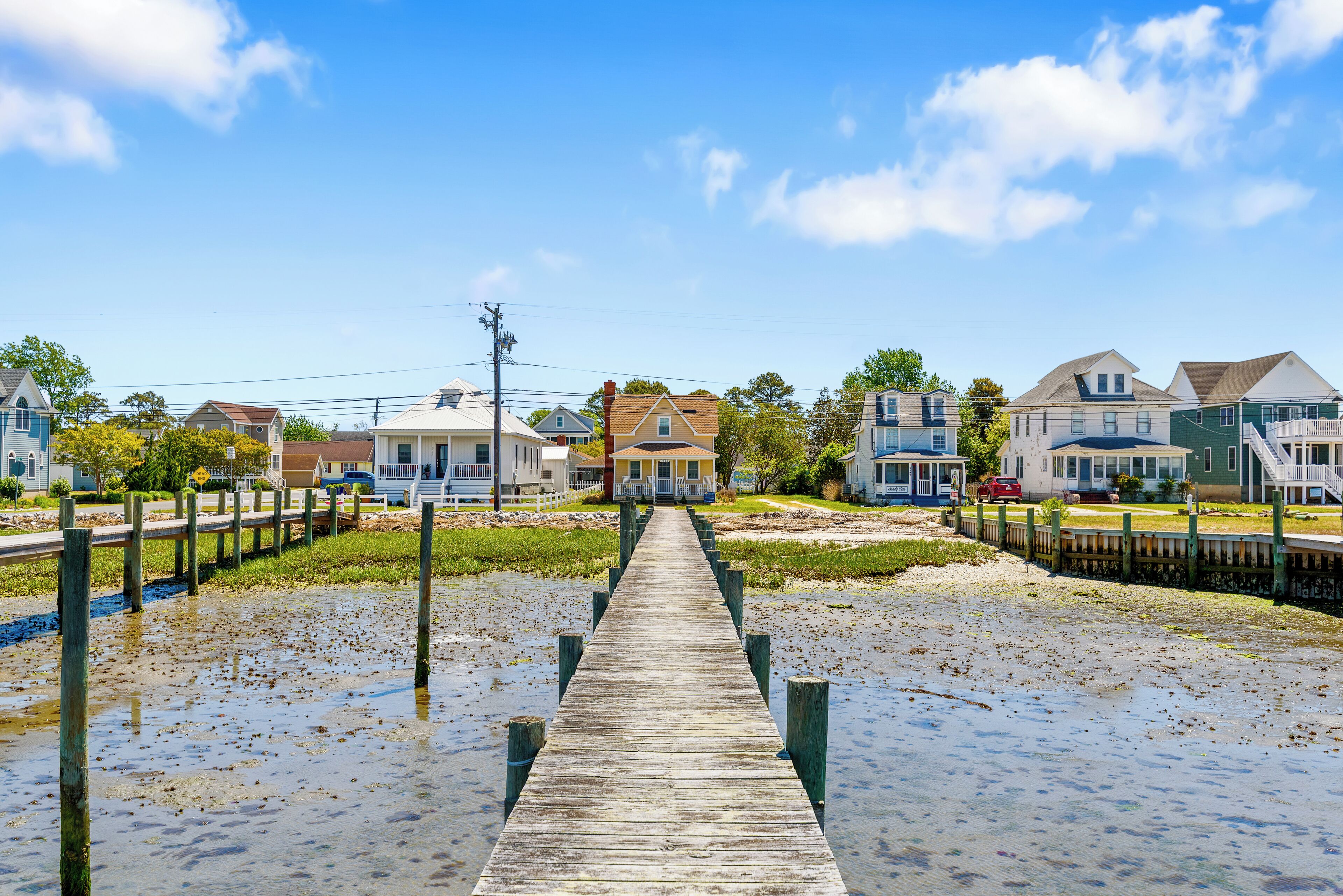 A wooden pier leading to a house with a porch, surrounded by other houses and a clear sky.