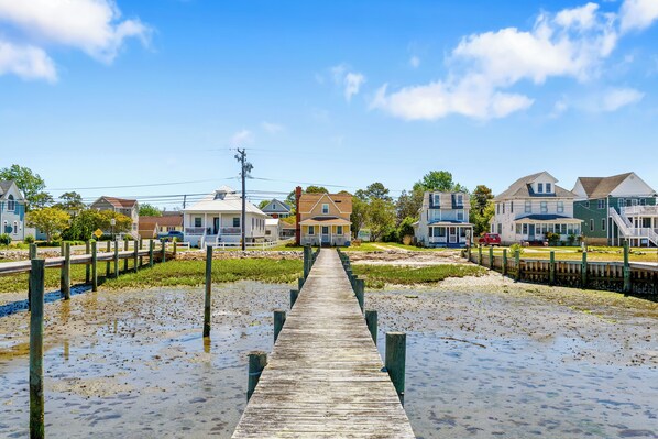 A wooden pier leading to a house with a porch, surrounded by other houses and a clear sky.