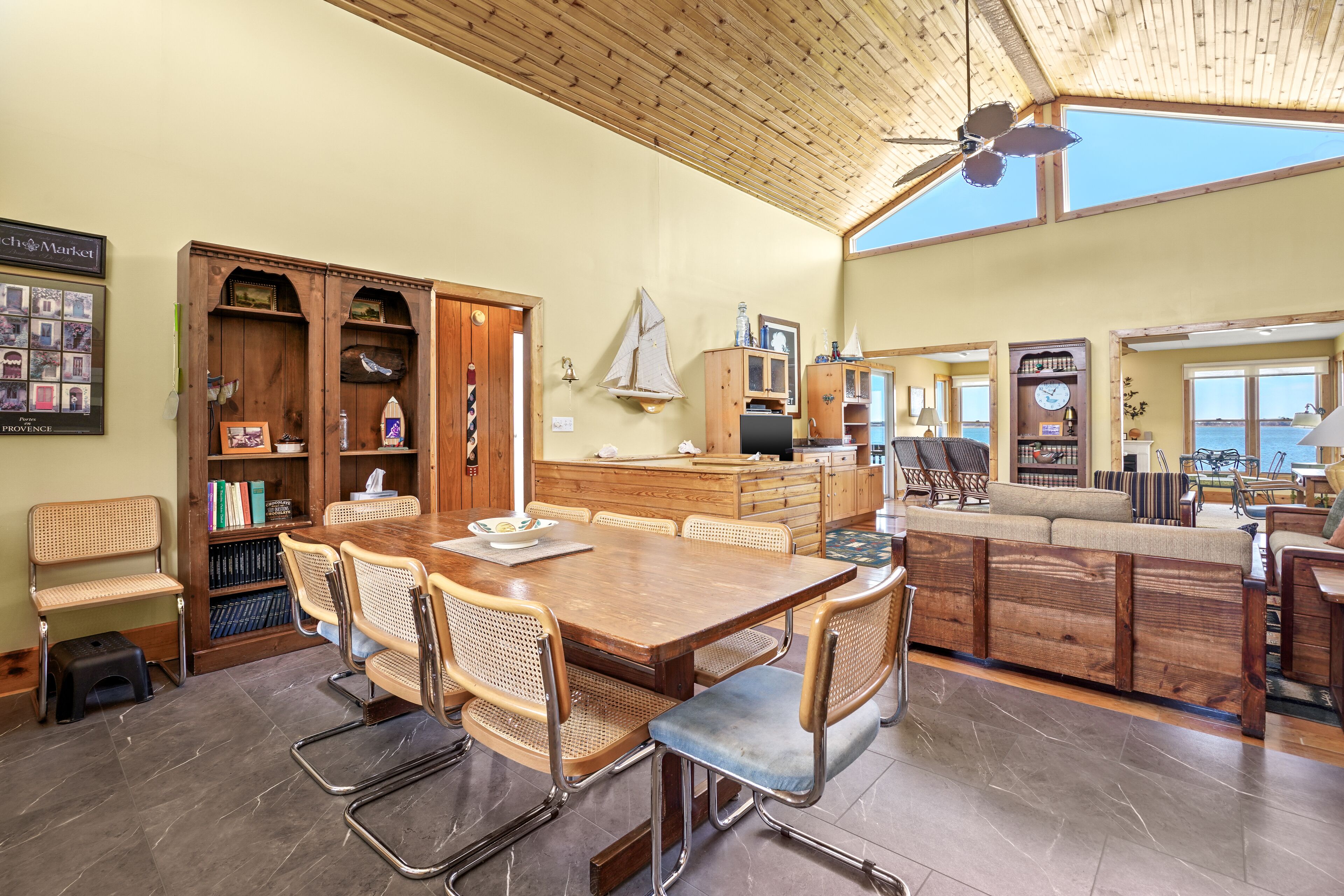 A spacious dining area with a wooden table and chairs, a wooden cabinet with books, and a view of the sea through large windows.