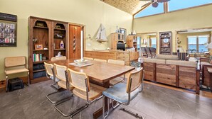 Un vaste espace dédié à la salle à manger, avec une table et des chaises en bois, un meuble en bois rempli de livres et une vue sur la mer grâce à de grandes fenêtres.