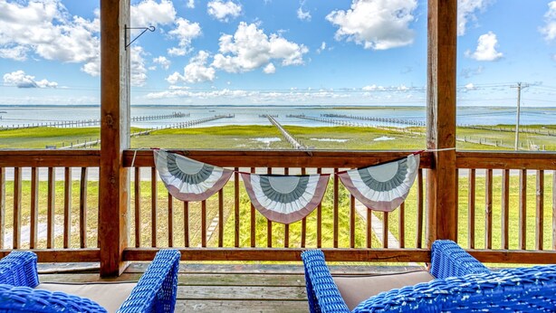 Une terrasse en bois avec des chaises en osier bleues, une vue sur une plage avec une jetée, et un ciel dégagé.