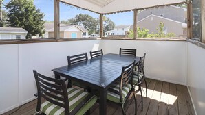 Un porche cubierto con una mesa y sillas de madera, ventanas amplias y vistas a casas vecinas.