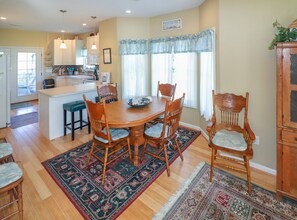 Uma sala de jantar com uma mesa e cadeiras de madeira, cozinha ao fundo e uma janela com cortinas.