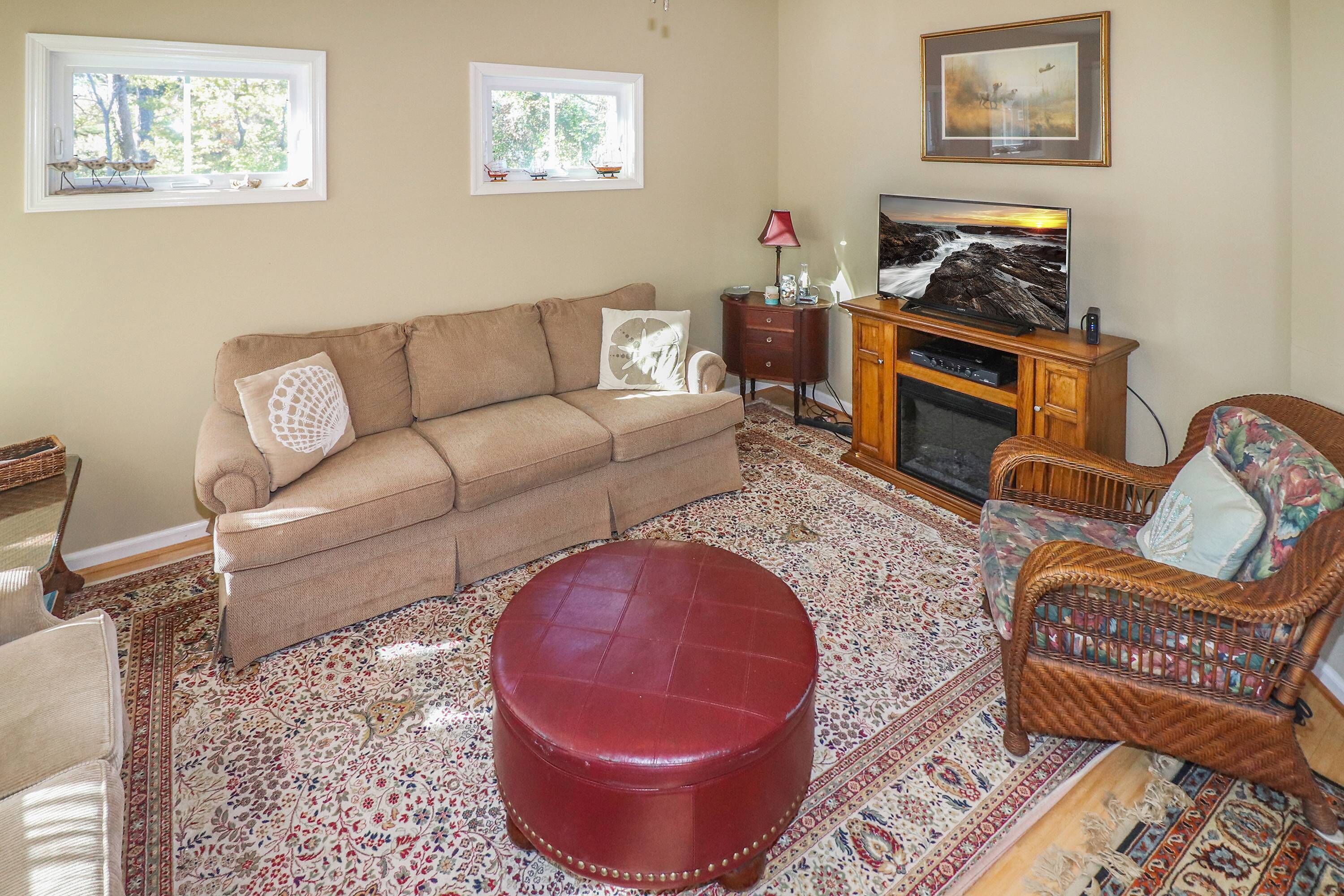 A living room with a beige sofa, a red ottoman, a wicker chair, a television, and a fireplace.