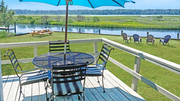 Une terrasse avec une table ronde et des chaises, un parasol bleu, et une vue sur un lac et la verdure environnante.