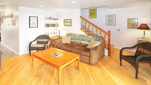 Un salon avec un plancher en bois, un canapé marron, une table basse en bois, des chaises en osier et un escalier.