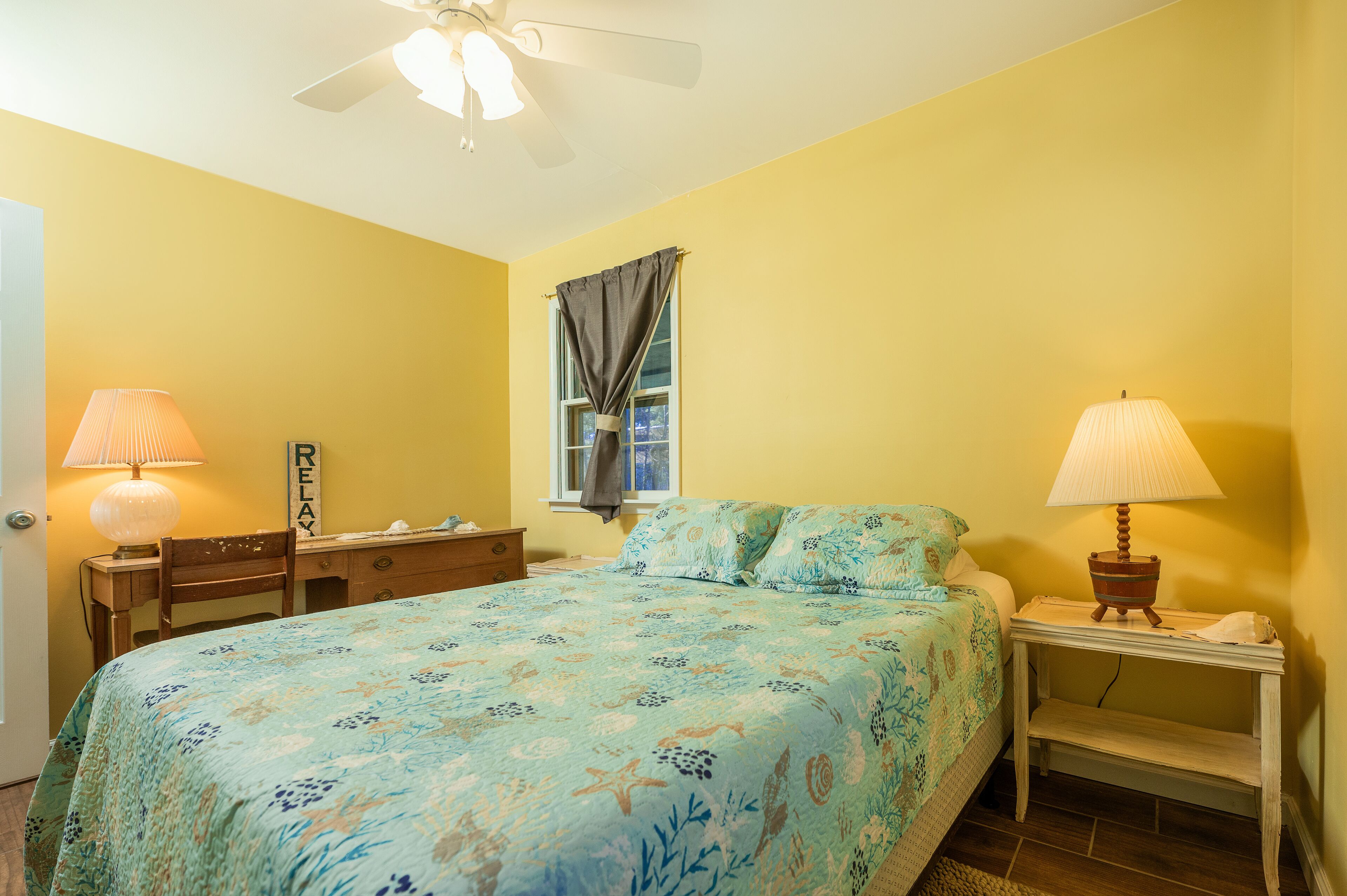 A bedroom with a bed, two bedside tables with lamps, a window with curtains, a ceiling fan, and a wall-mounted sign.