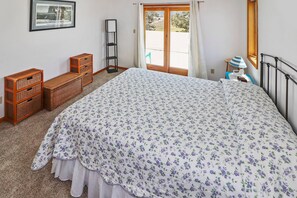 A bedroom with a large bed, a wooden bench, wicker baskets, a framed picture on the wall, and a window with curtains. - Herbie's Hideaway | Luxury Water Views, Dock Access & Screened Porch Near Memorial Park (Chincoteague Island)