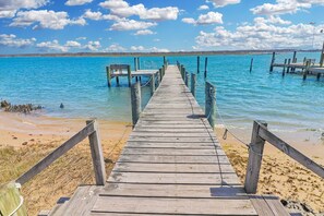 A wooden pier leading to a beach with clear blue water and distant land. - Herbie's Hideaway | Luxury Water Views, Dock Access & Screened Porch Near Memorial Park (Chincoteague Island)
