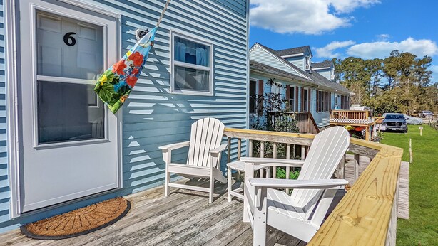 A deck with white chairs and a table, a hammock, and a house with blue siding and a number '6' on the door. - Heron Landing | Remodeled Coastal Cottage, Walk/Bike to Memorial Park (Chincoteague Island)