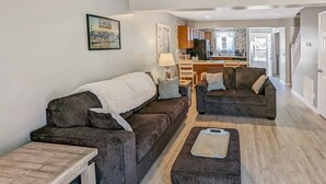 A modern living room with dark sofas, a wooden coffee table, and a dining area with wooden cabinets and a kitchen in the background. - Heron Landing | Remodeled Coastal Cottage, Walk/Bike to Memorial Park (Chincoteague Island)