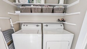 A laundry area with two white front-loading washing machines, a drying rack, and shelves above holding baskets and cleaning supplies. - Heron Landing | Remodeled Coastal Cottage, Walk/Bike to Memorial Park (Chincoteague Island)