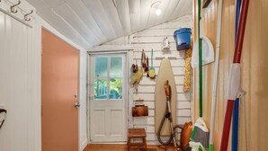 A hallway with a wooden door, a small wooden stool, a surfboard, and cleaning tools. - Homestead | Historic Cottage on 30 Acres, Porches, Bay Breezes, Outdoor Shower, Nature Retreat (Chincoteague Island)