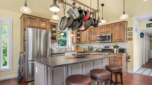 A modern kitchen with stainless steel appliances, wooden cabinets, and a hanging pot rack. - Homestead | Historic Cottage on 30 Acres, Porches, Bay Breezes, Outdoor Shower, Nature Retreat (Chincoteague Island)