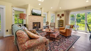 A spacious living room with a fireplace, comfortable seating, and a large window offering a view of the outdoors. - Homestead | Historic Cottage on 30 Acres, Porches, Bay Breezes, Outdoor Shower, Nature Retreat (Chincoteague Island)
