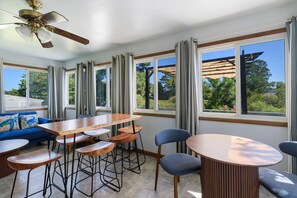 A modern dining area with a wooden table and chairs, large windows with curtains, and a ceiling fan.