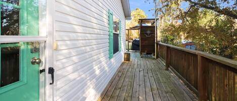 Une terrasse en bois, avec une maison blanche dotée de volets verts et d’un petit porche.