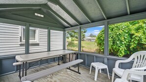 A covered outdoor area with a table and chairs, a bench, and a view of greenery and a house. - Lazy Days | Main Street Cottage w/ Snippet Bay Views, Close to Town + Trails, Pet Friendly (Chincoteague Island)