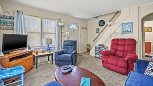 A living room with a television, a blue armchair, a red armchair, a wooden coffee table, and a bookshelf. - Lazy Days | Main Street Cottage w/ Snippet Bay Views, Close to Town + Trails, Pet Friendly (Chincoteague Island)