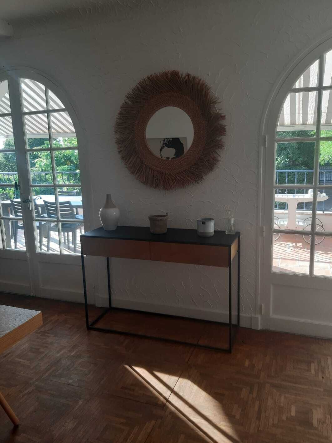 A room with a wooden floor, a round mirror on the wall, a small table with a vase and a candle, and large windows with a view of outdoor furniture.