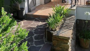 Een pad van stenen leidt naar een terras met buitenmeubilair, potplanten en een gebouw met boogvensters en een betegeld dak.