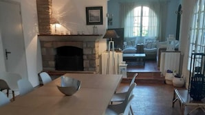 Een ruime eethoek met een lange houten tafel en moderne stoelen, een stenen open haard en een gezellige zithoek met een bank en grote ramen.
