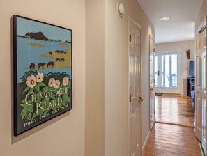 A hallway with a framed poster of Chincoteague Island, a door, and a staircase leading to another room. - Marsh Point | Marsh Views + Shared Dock, Screened Porch Dining, Cozy Island Townhome (Chincoteague Island)