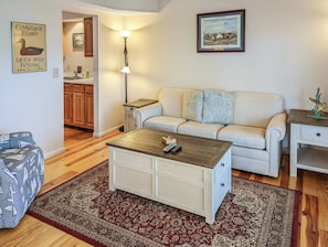 Uma sala de estar com sofá, mesa de centro, tapete, abajur e um quadro na parede.