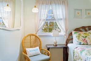 Een slaapkamer met een rieten stoel, een nachtkastje met een lamp, een bed met een bloemenprint sprei en een raam met dunne gordijnen.