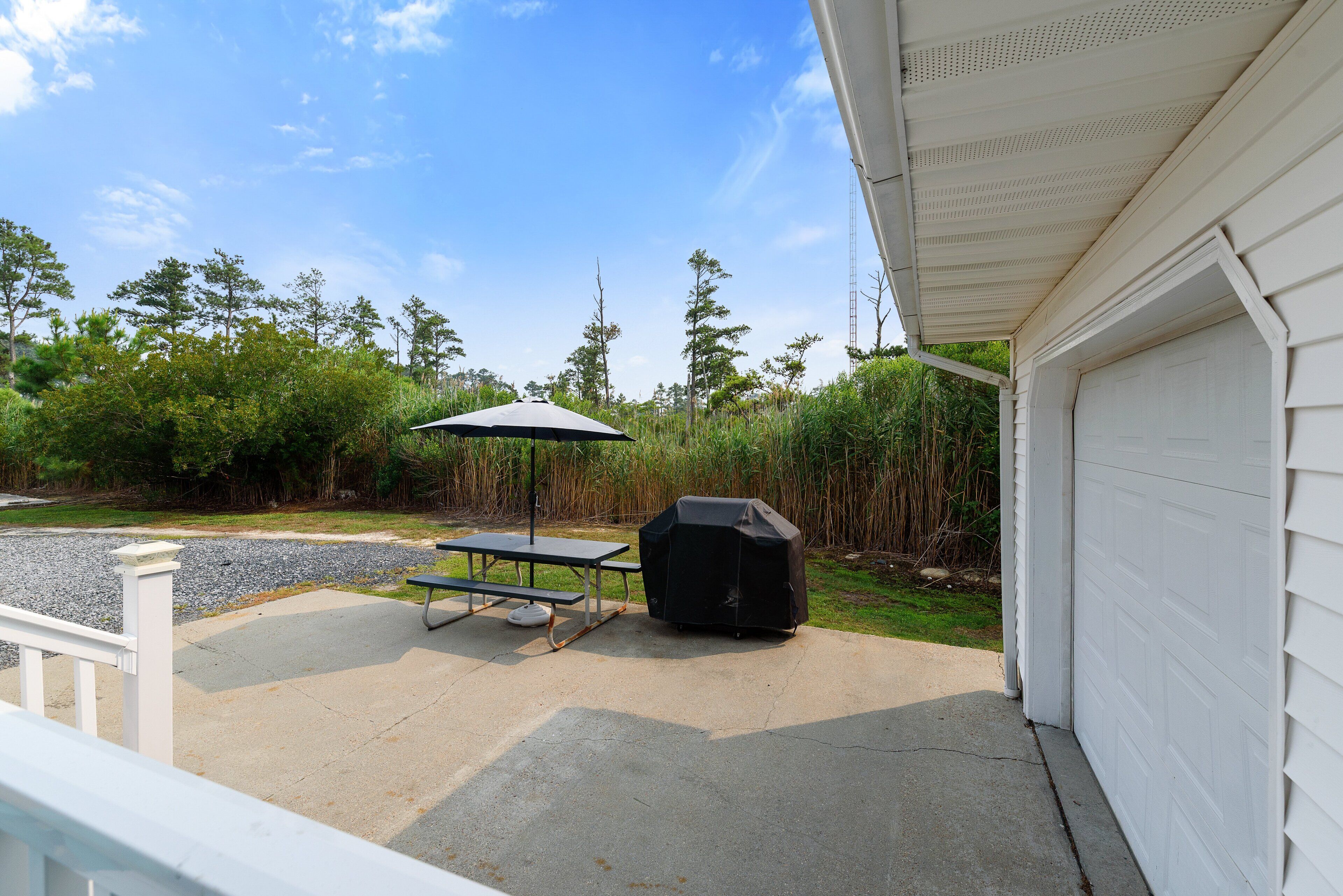 A patio with a picnic table and grill, surrounded by vegetation and a clear sky.